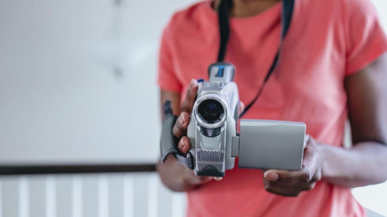 Stock shot of torso of anonymous black young person holding a video camera pointing directly to camera