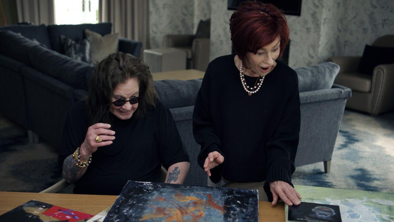 A man and woman are both wearing black and looking shocked at paintings on a table.