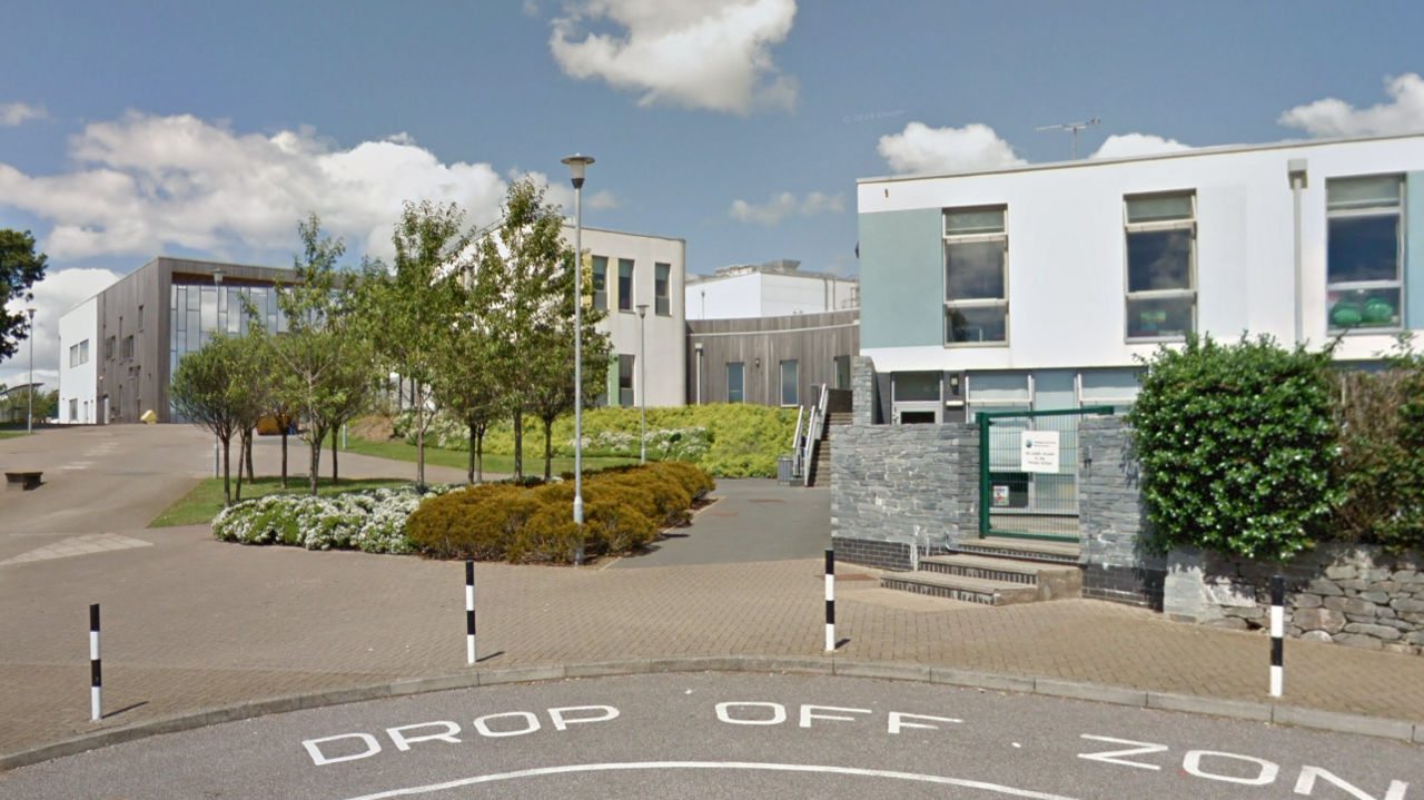 A street view of Sir John Hunt Community Sports College. There is a white building in the foreground and in the background are more college buildings. Street paint at the forefront of the images shows a designated 'drop off zone' for the school.