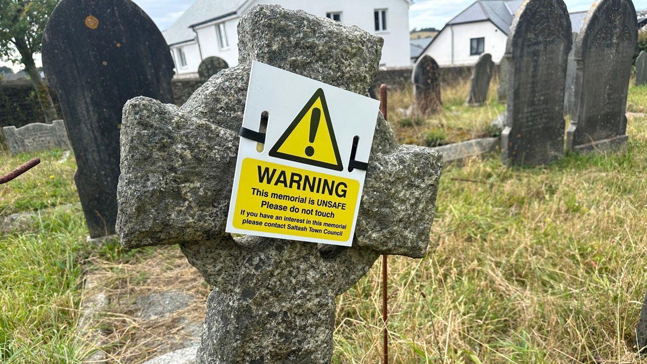 A headstone in the shape of a cross. It has a warning sign on it. It is tilted slightly to the right. The grass is green and brown. There are headstones in the background.