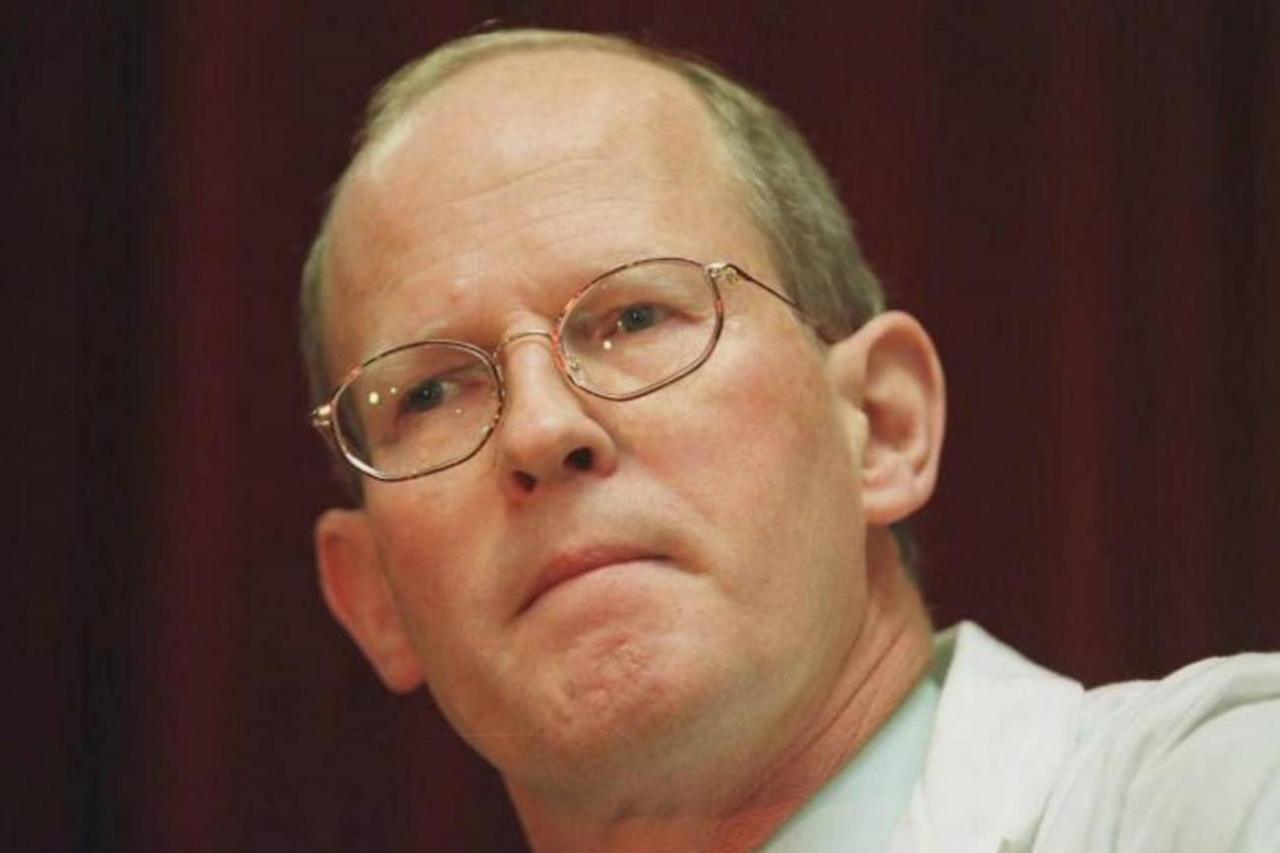 A man with thin-framed glasses and short mousy brown hair. The picture looks old due to its quality. It is a head and shoulders shot. He has a white lab coat on.