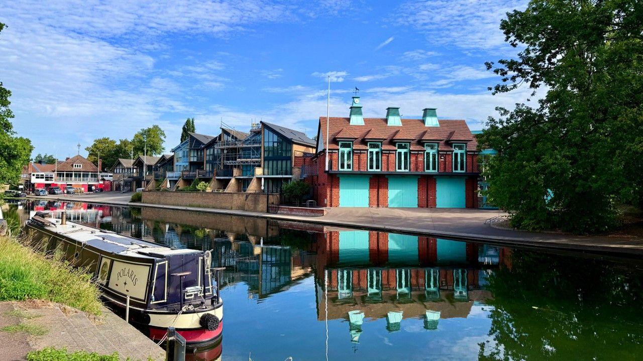 A blue and cream barge moored on the side of a canal and buildings on the other side, with blue sky and broken cloud above