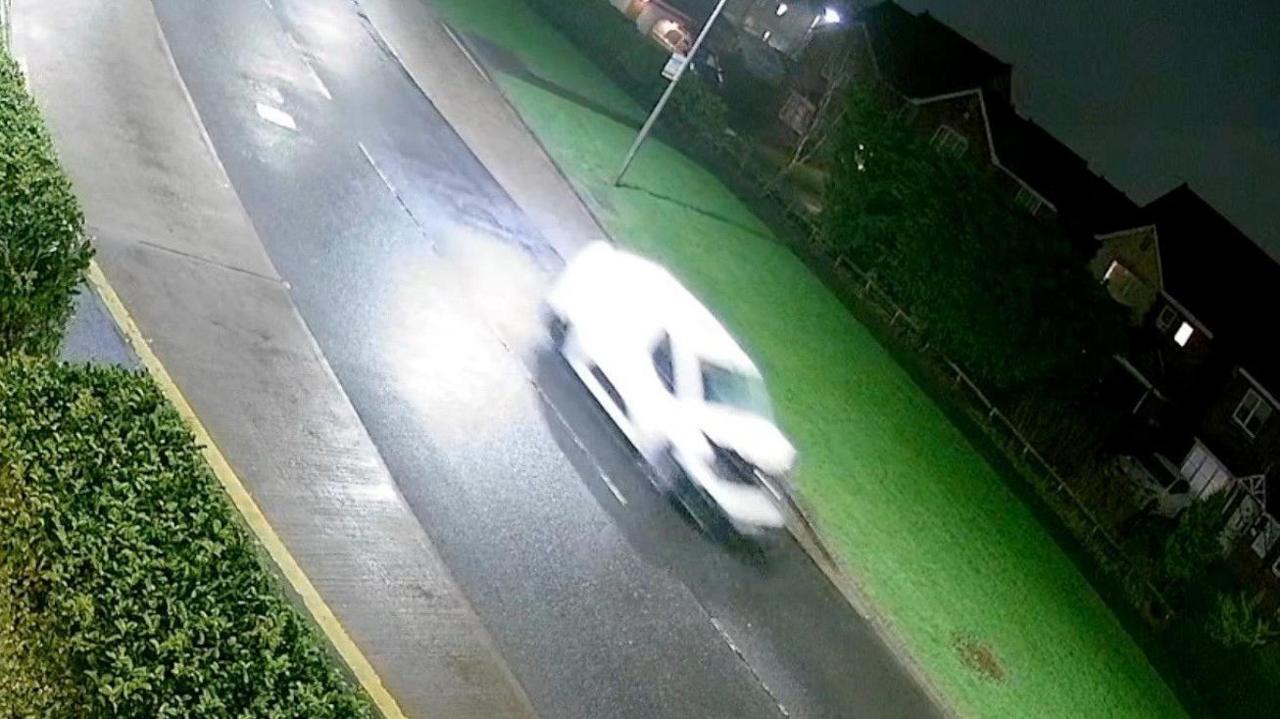 Still from a CCTV footage of a white van driving on a road past houses, its windscreen is smashed and bonnet is crumpled upwards