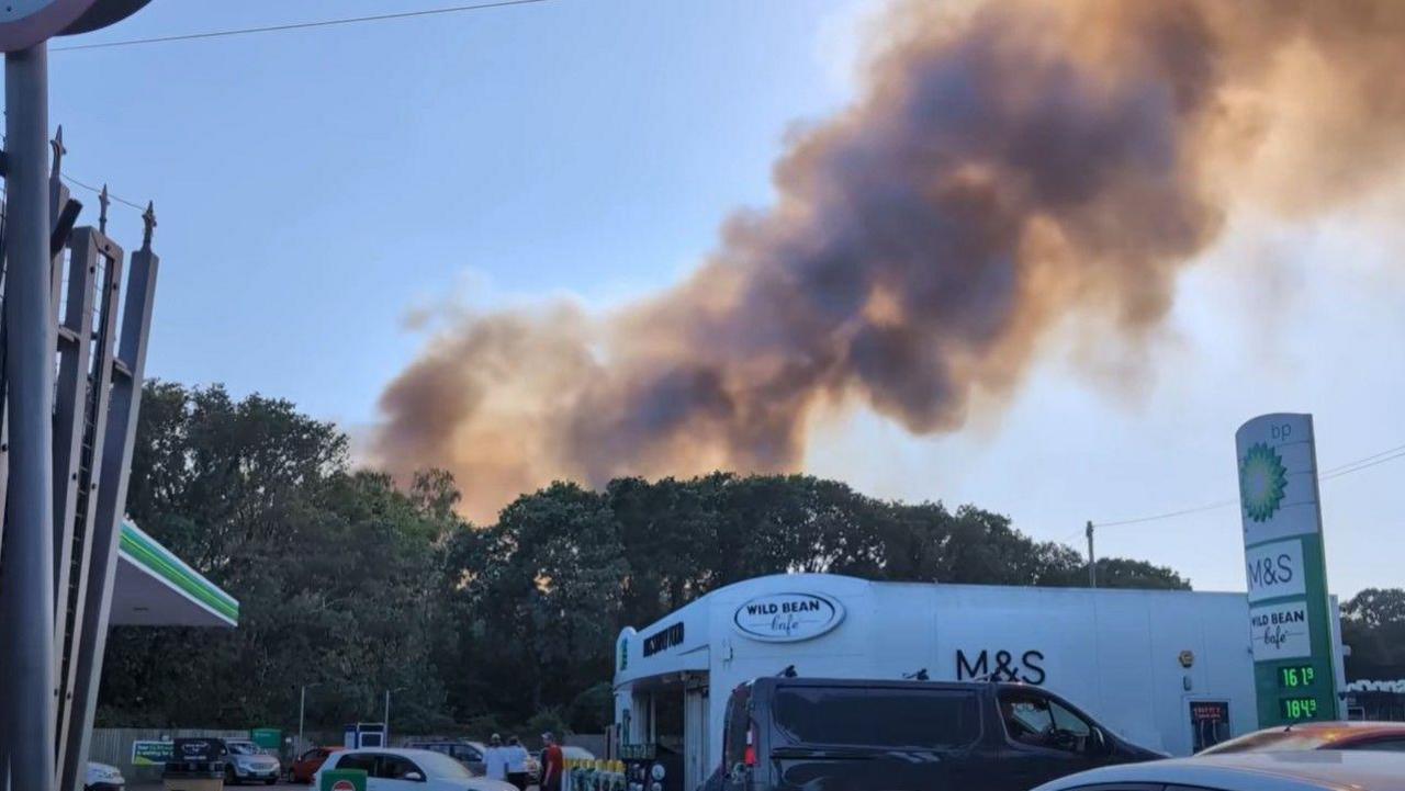 Black and orange smoke billow from behind tall trees which are in front of a petrol station with cars moving on the forecourt.