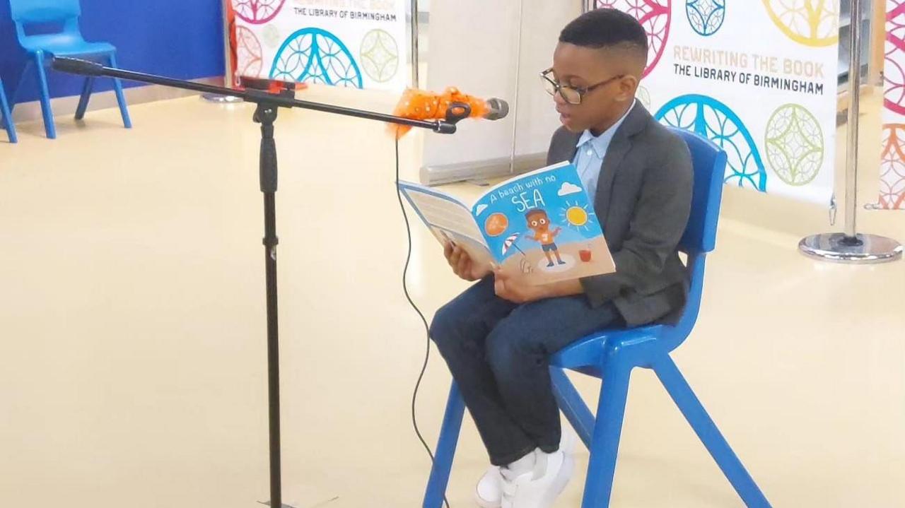 Jayce sitting on a blue chair with his book A Beach With No Sea as he reads into a microphone that has been set up in front of him.