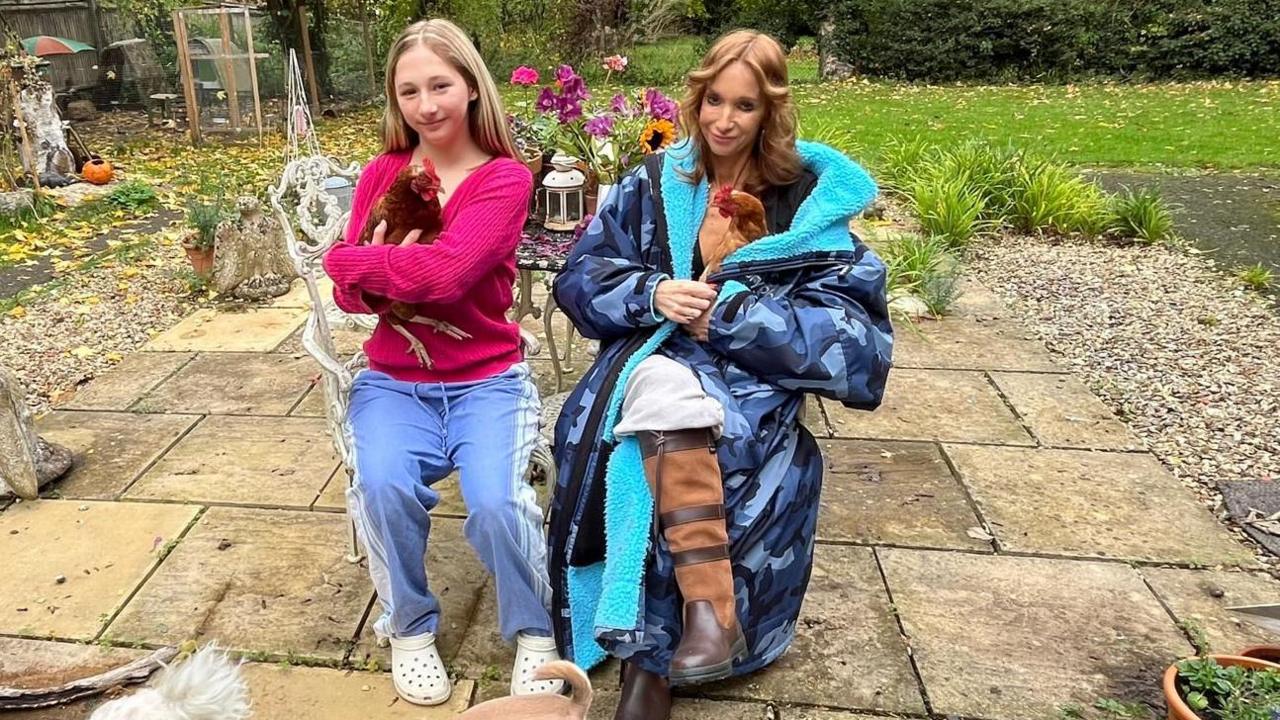 Victoria is sitting on a chair in a garden. She is wearing a blue coat and brown boots and is holding a chicken. Her daughter Poppy is standing next to her and is holding another hen. Two dogs are in front of them.