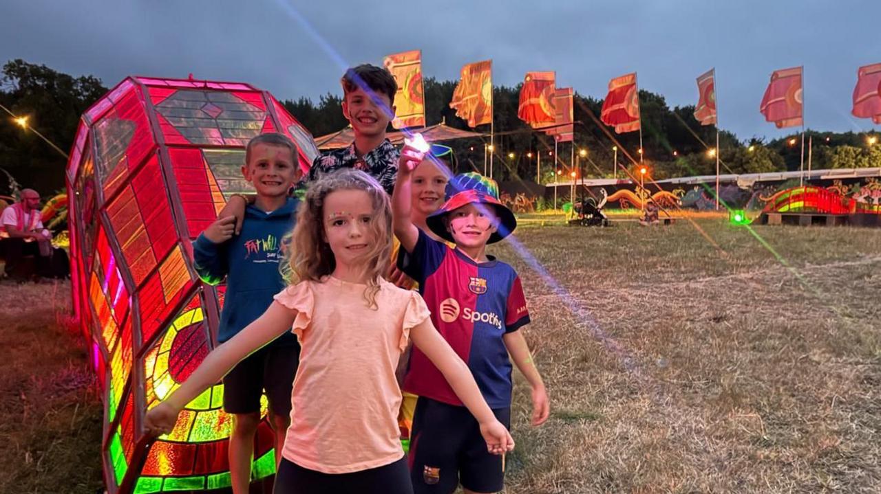 Five children stand next to Glastonbury's Dragon's Tail - a 40m long art installation of stained glass. The children are next to the Dragon's head which is lit up in red, yellow and green.