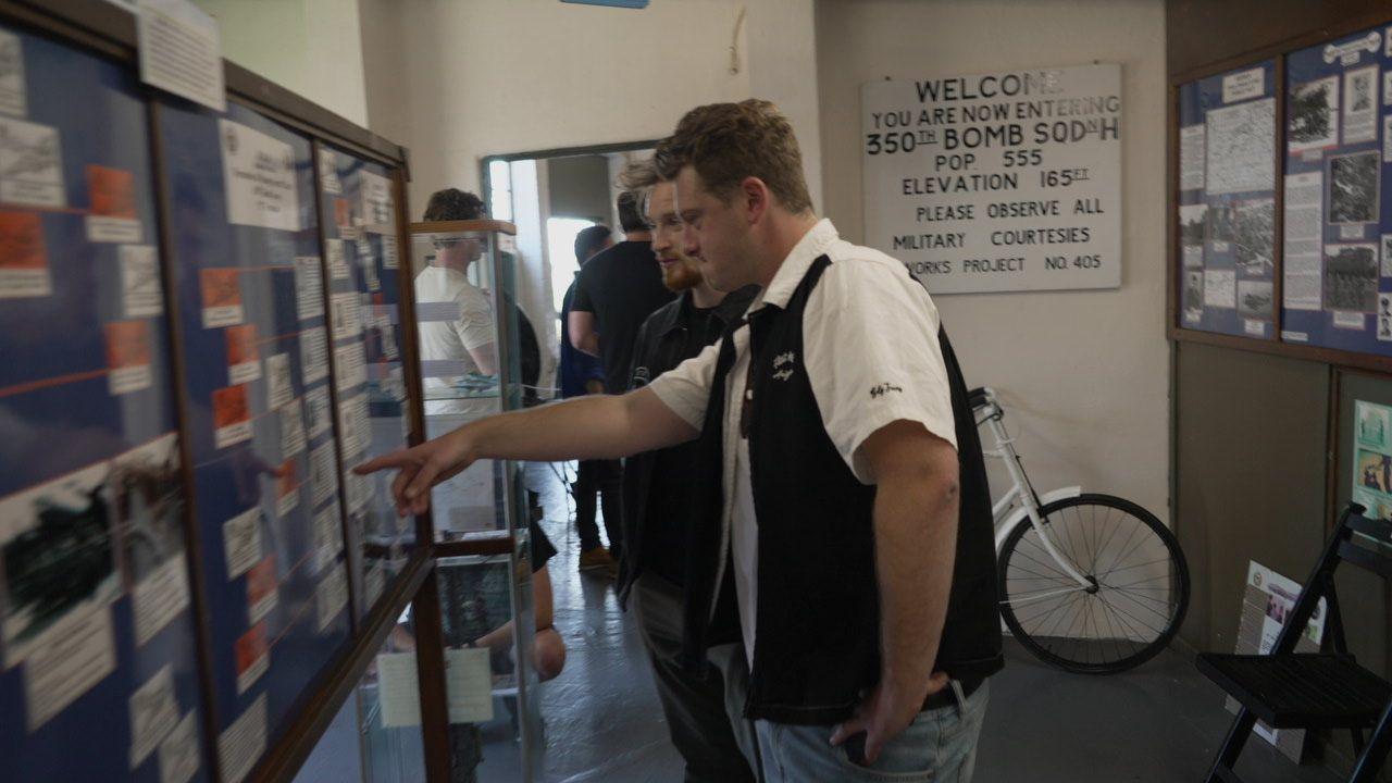 Actor Josh Bolt is wearing a white shirt and black sleeveless jacket and standing inside the museum, pointing at an artefact on the wall.