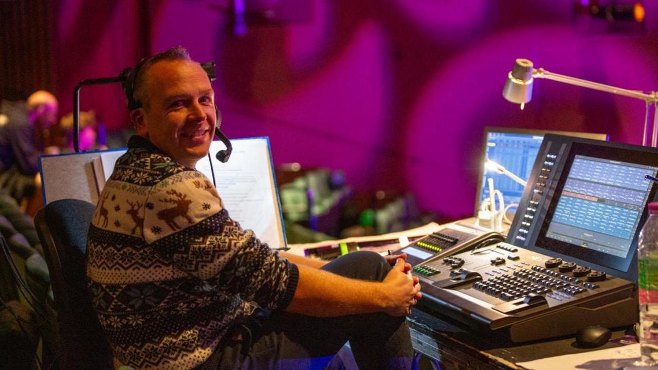 Jonathan Haynes wearing a festive jumper sits at a theatre control desk with colourful lighting and screens in the background, smiling towards the camera.