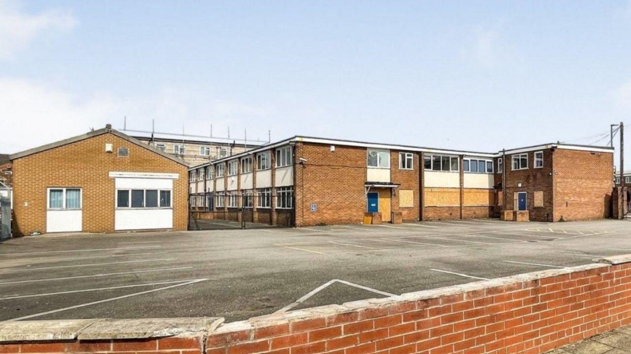 A car park and side view of a large, two-storey bricked building which is partially boarded up. 