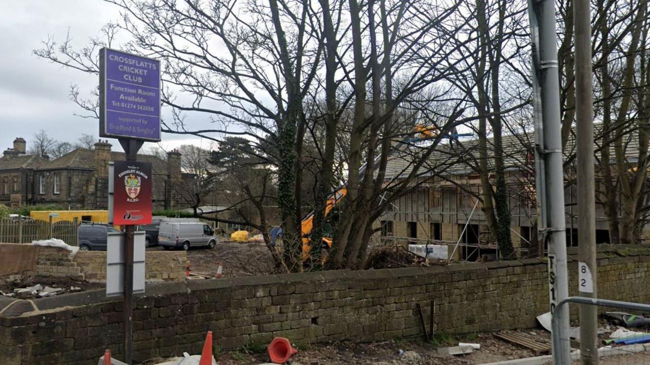 A tall blue signpost bearing the name Crossflatts Cricket Club in front of a building with scaffolding around it