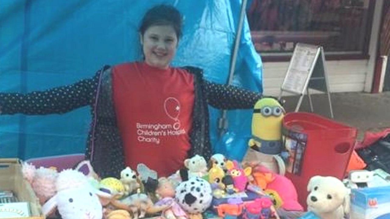 A photo of Ms Giorgio-Jones at a younger age than now, smiling and wearing a mainly red Birmingham Children's Hospital Charity T-shirt. She is standing behind a table containing soft toys. 