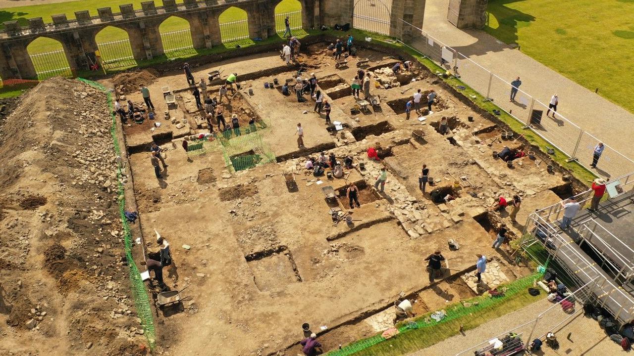 An aerial shot of archaeologists digging at Auckland Castle in the sunshine. The large site is fenced off. 