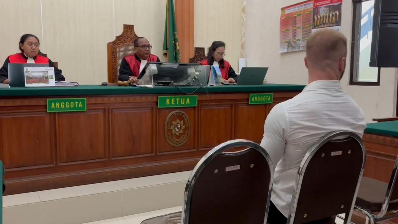 A picture of the Bali court room. The picture is taken behind Mr Parker who is sitting in front of three judges. The judges are wearing black shirts and red jackets with no sleeves. They are sitting at a large wooden desk.