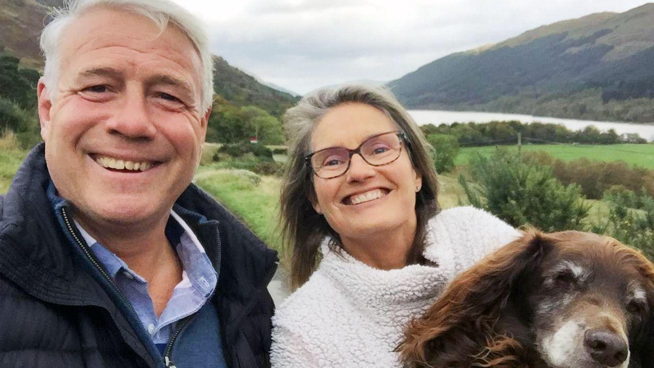 A selfie of Scott and Jenny Hastings with their brown spaniel, standing outdoors with a valley in the backdrop