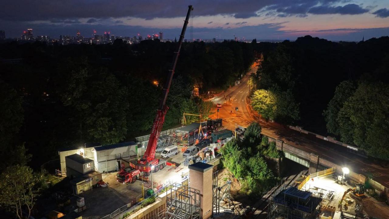 An aerial view of a construction site at night, with lights illuminating a large red crane which is holding a large black metal object and a road is seen in the distance