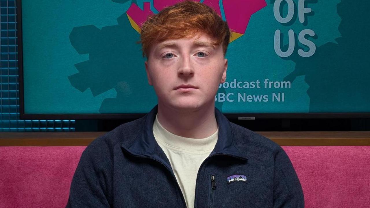 A young man with red hair looks into the camera with a neutral impression on his face. He is wearing a white t-shirt and a blue zip-up fleece jacket. He is sitting in The State of Us podcast studio, which has pink upholstered sofas and a blue walls. Behind his head is a TV screen with The State of Us logo - a pink rendering of the map of Northern Ireland along with the words The State of Us 