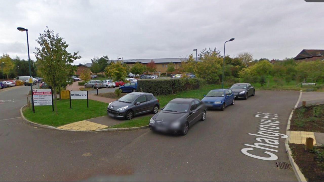 A parking area surrounded by trees and bushes. A building is visible in the background. Nearby sign reads "OAKHILL STC"


