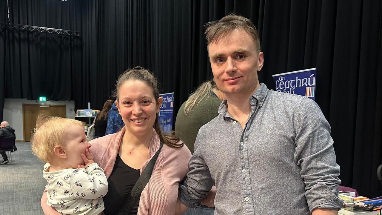 A woman and man standing together, while she holds a baby. She has a dark ponytail, pink cardigan and black top. The man has short dark hair and is wearing a grey shirt.