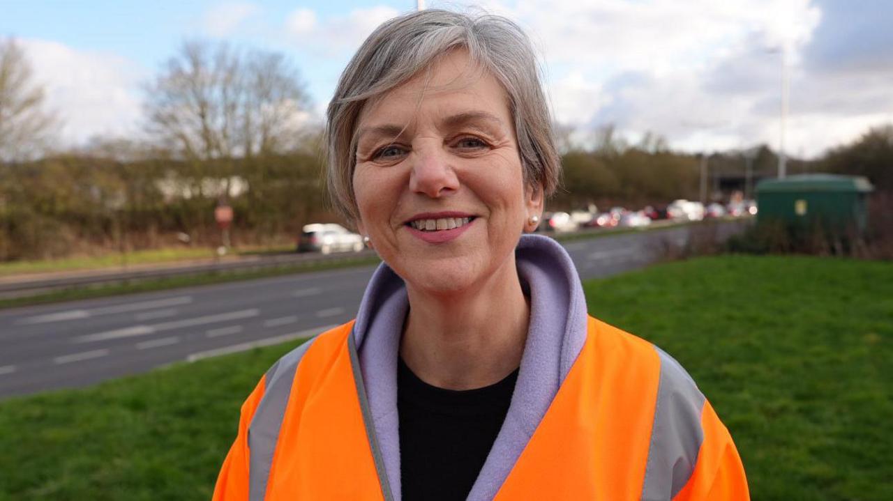 A woman wearing an orange high vis jacket standing on a grassy area next to a dual carriageway.