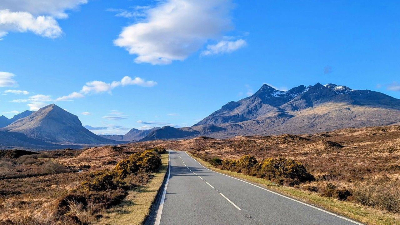 An empty road extends into the distance with brown shrubs on either side and blue, brown mountains in the background and blue, sunny skies above