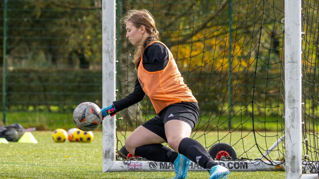 A goal keeper on the team, who has an amputated arm, catches a ball with her hand. She is bending down to catch the ball while in goal. She wears an orange vest over a black t-shirt, as well as black shorts, black socks, and blue trainers.