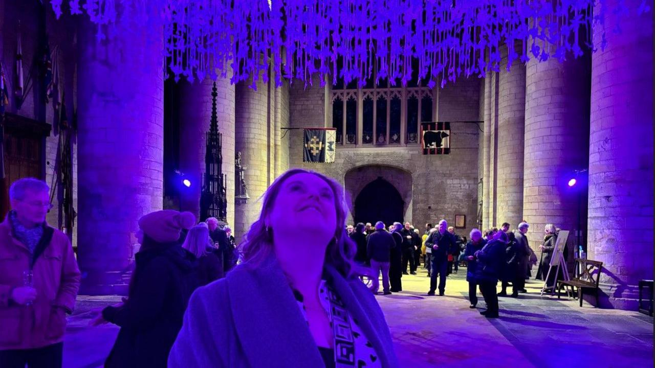 Thousands of paper doves hung from the ceiling of an abbey with purple lighting shining over them and a person standing beneath them looking up at them