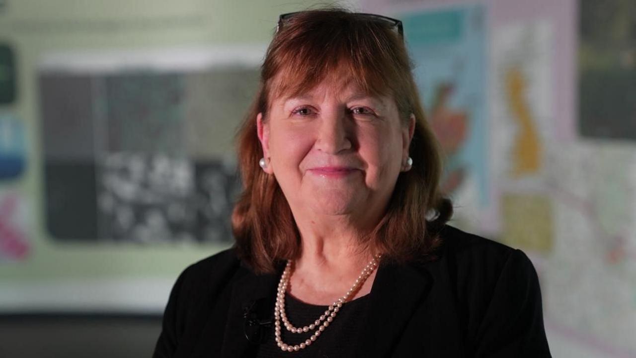 Professor Dame Lorna Dawson, a mature lady with auburn hair, glasses on her head and wearing pearl earrings and a pearl necklace smiles at the camera. Behind her on the wall we see a map.