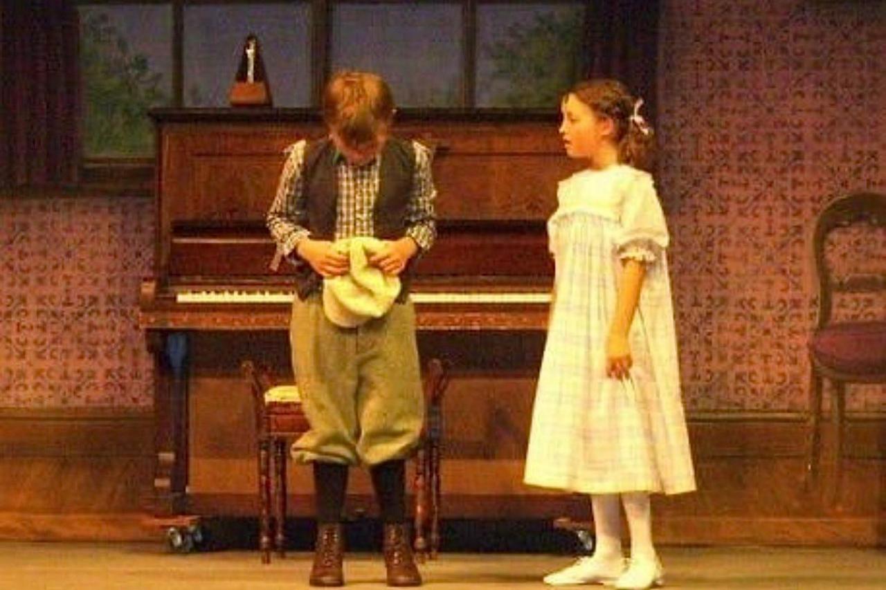 Two young actors are on stage in front of a piano. Bella Ramsey is on the right wearing a dress and a boy on the left has his head down and is holing a cap.