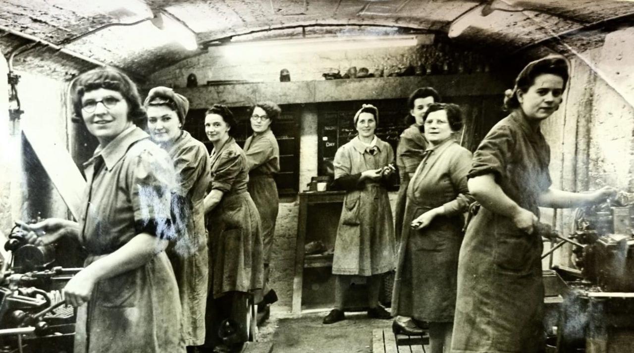 A group of women work in a makeshift munitions factory beneath the arcade in New Brighton, Merseyside. They are dressed in work overcoats and have their hair tied up or under headscarves as they operate machinery making bullets and shell casings.