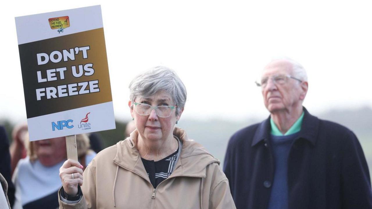 An elderly woman holds up a sign saying 'Don't let us freeze. She has grey hair and a beige coat.