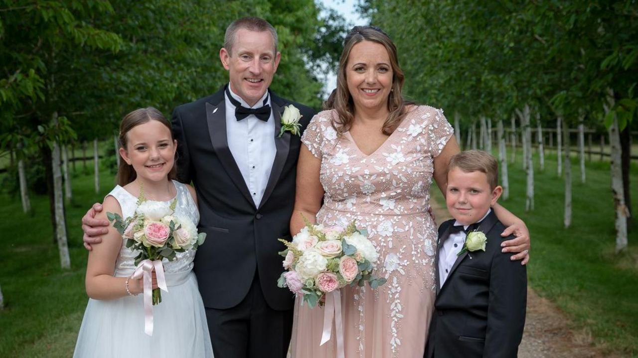 A photo with a family of four. From left to right there is a young girl in a white bridesmaids dress holding a bouquet of flowers. A father wearing a fitted tux and bow tie. A mother wearing a blush pink wedding dress and a young boy in a black suit and tie.