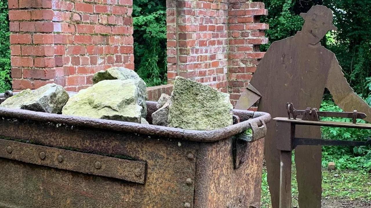 An old mine cart is full of green/grey lime stone. beside it is a metal cut-out sculpture of a man.