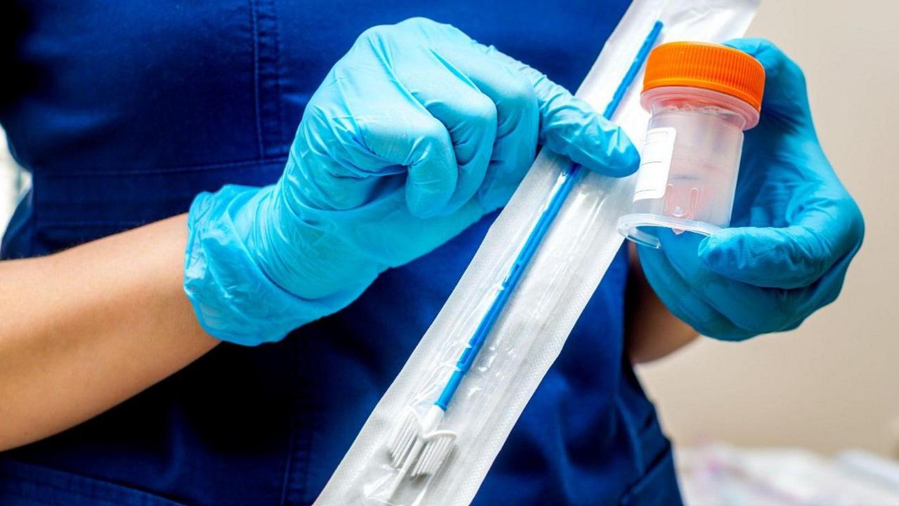 Person with blue scrubs and latex gloves holding a swab and sample cup.