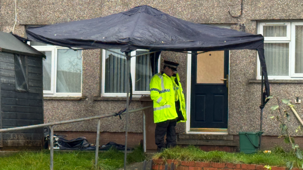 A police officer stands by the door of a home, with a forensic tent visible