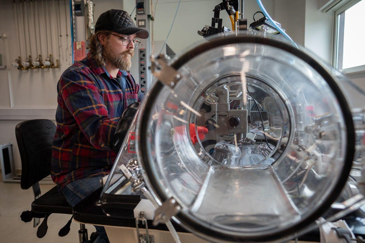 James Malley sits on a chair with his hands inside a long see-through tube. Inside are wires and metal pieces of equipment.