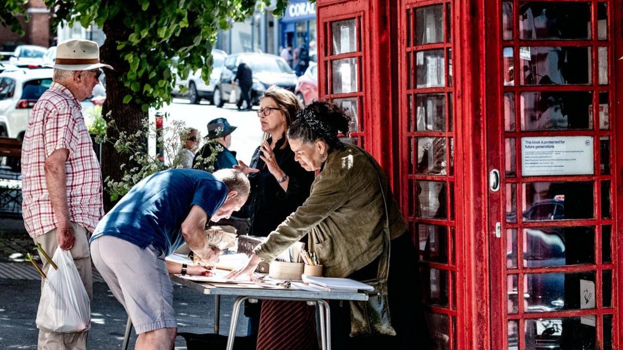 Lisa Wilson and Kim Squirrell standing behind a table next to the two red phone boxes. They are speaking to two men. One of them is bending over the table and writing.