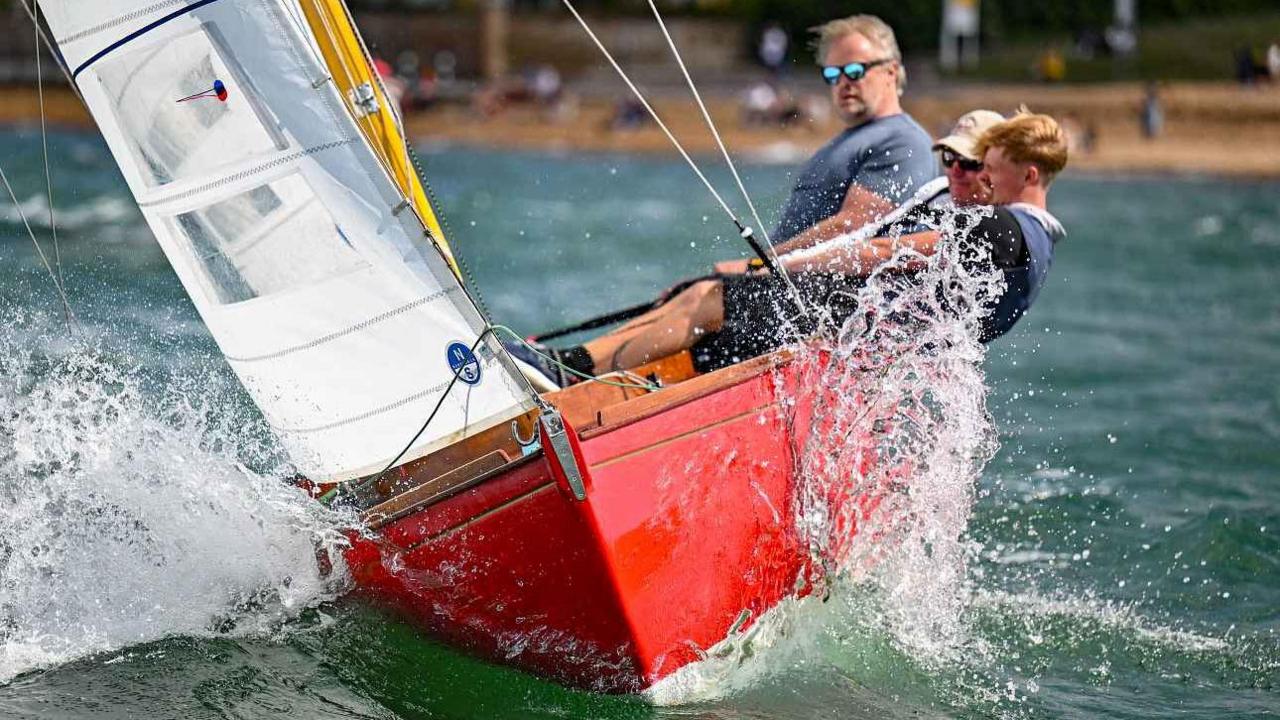 Three people sailing a Magic Dragon XOD class sailing boat. The boat is red with a white sail and the people are leaning back as the boat tilts.