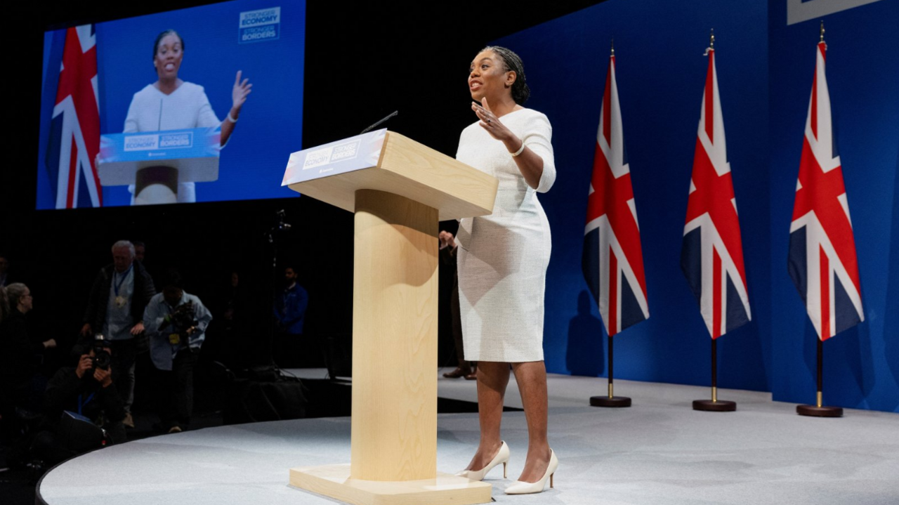 A woman in a white knee-high dress with white heels stands at a wooden lectern with a large screen behind her and three union jack flags.