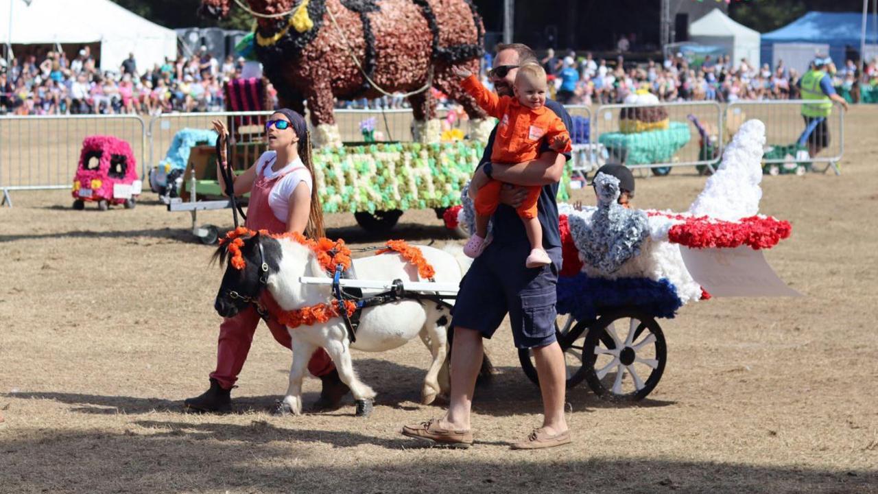 A man and woman take their flower display across the showground during the North Show's Battle of Flowers. Their display has a miniature white pony pulling it. The man is carrying a baby who has an orange jumpsuit on.