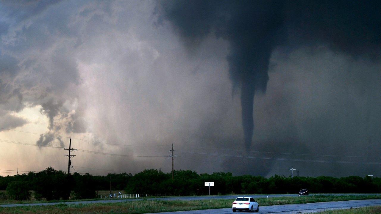 A huge tornado sweeps across the ground with a motorway and car in the foreground.