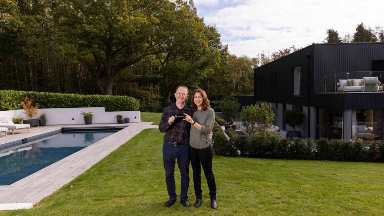 Richard and Yannick Baker stand in front of the house next to the swimming pool. Behind them are big trees. Richard is wearing a checked red and navy shirt with blue skinny jeans. He is wearing glasses and has short hair. Yannick is wearing black skinny jeans and is wearing a taupe shirt. She has long brown hair.