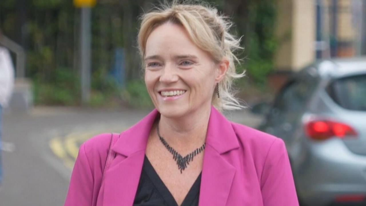 Sandie Peggie - a woman with short blonde hair and wearing a pink jacket and dark top - smiles outside a tribunal hearing.