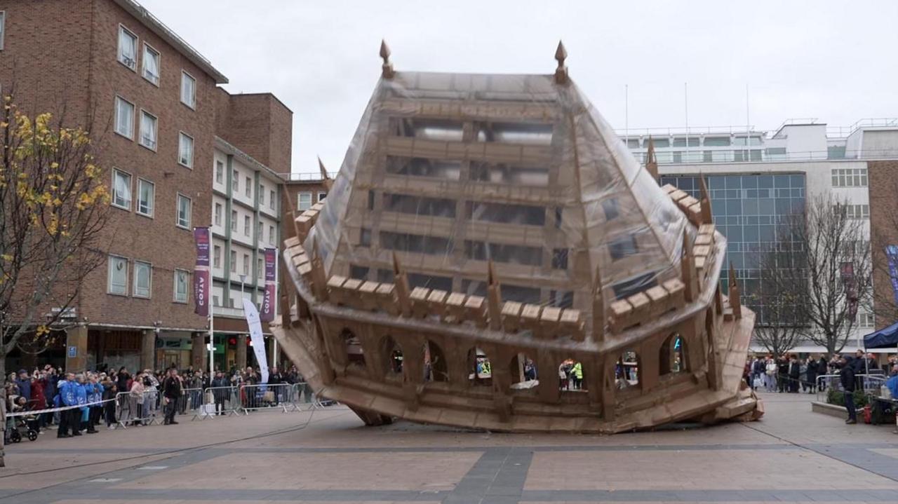 An art installation made of cardboard representing Coventry Cathedral. It is 15ft high and in a public square, surrounded by shops and members of the public. It is lying partially on its side. Crowds behind a barriers surround it.