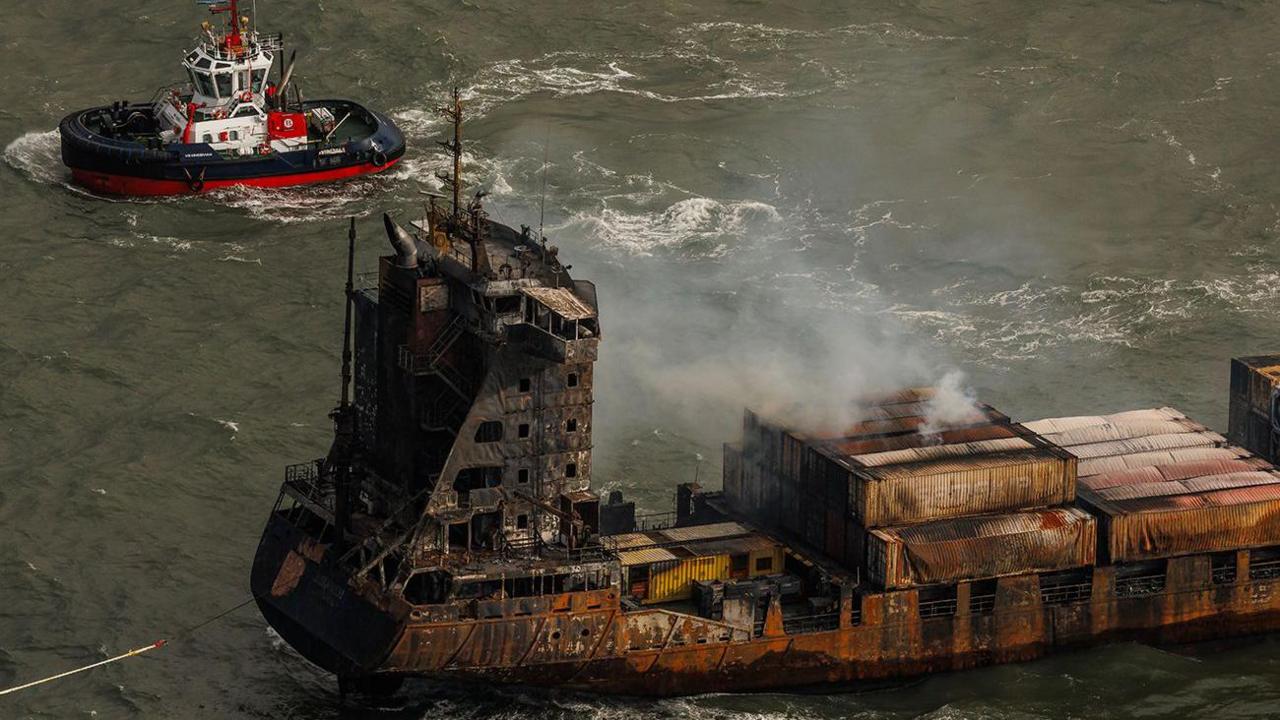 Smoke billowing from the fire-damaged Solong cargo vessel in the sea. A small tug boat is nearby on the top left.