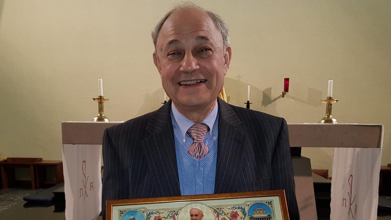 Edgar Riches in a dark jacket with light blue shirt and jumper, standing in front of a church altar.