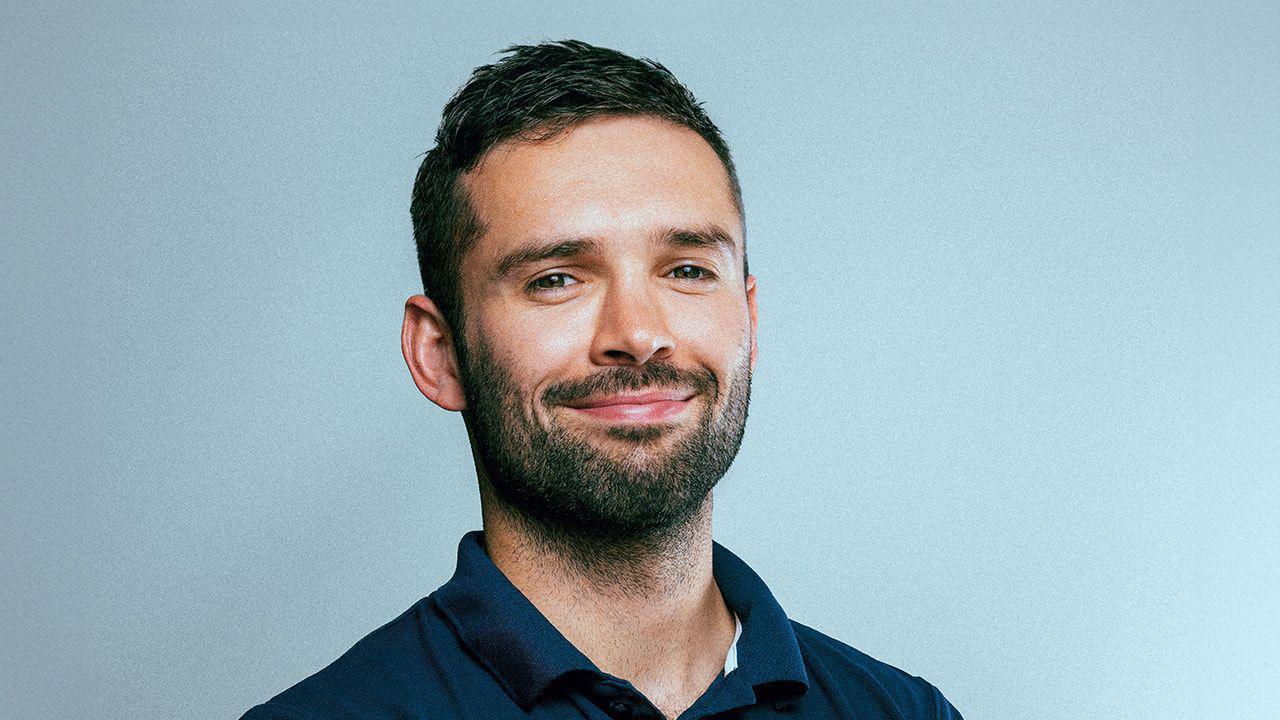 Ted Morgan smiles at the camera. He has short dark hair and a dark beard. He wears a navy polo t-shirt and stands against a light blue background. 