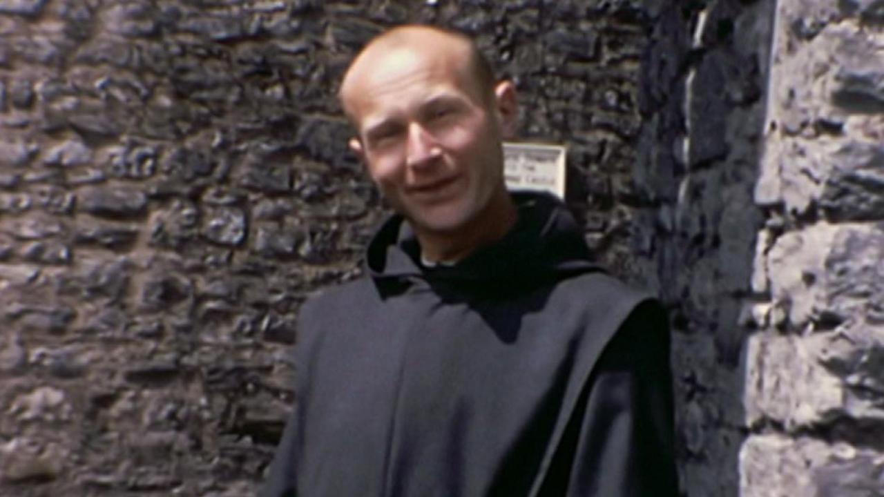 Father Thaddeus Kotik stands in front of the walls of Pembroke Castle. He is wearing a black habit. He is bald, with a little dark hair on the sides of his head. He is smiling into camera. 