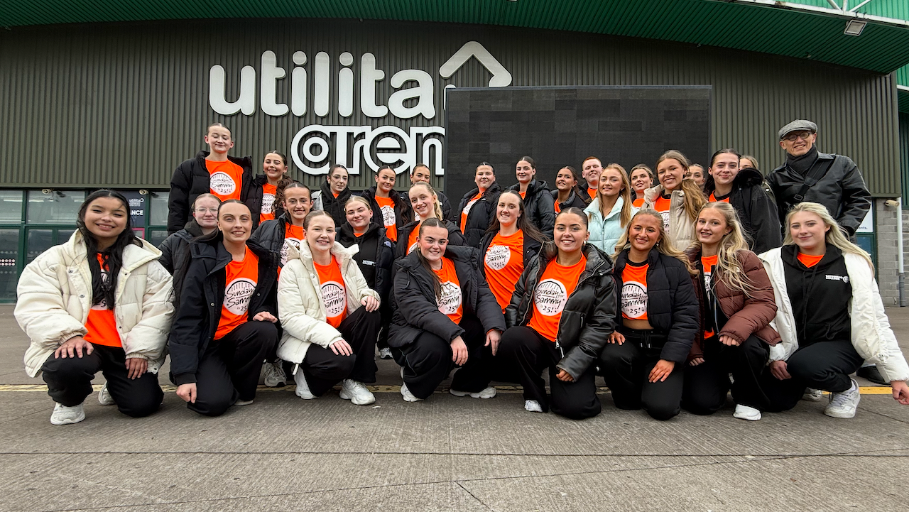 A group of students wearing a orange T-shirts and black trousers, stand in front of a dark green-coloured arena entrance.