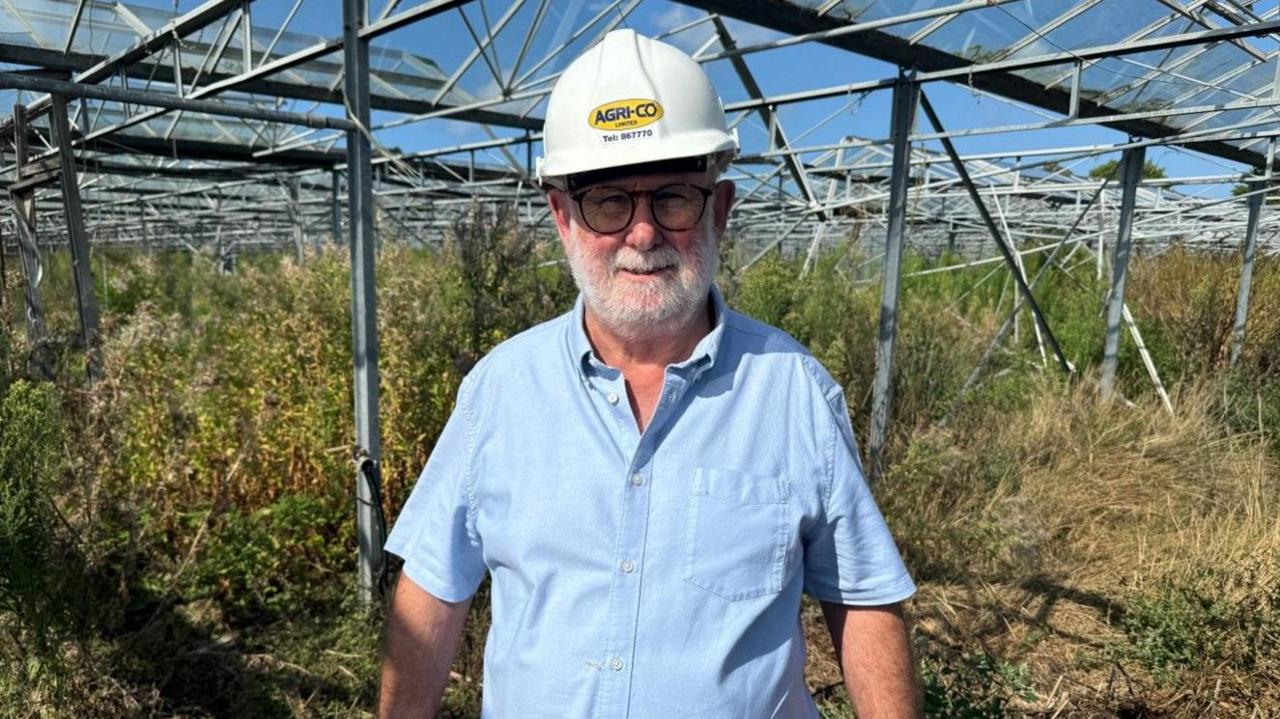 Kevin Hervé, in a blue short-sleeved shirt and white hard hat, standing inside a redundant glasshouse in which vegetation has grown to waist height.